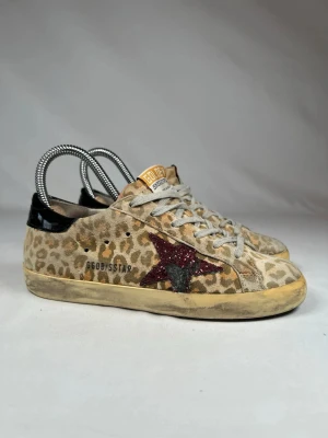 Leopardmönstrade Golden Goose sneakers - Snygga sneakers från Golden Goose med leopardmönster i beige och brunt, rödglittrig stjärna på sidan och svarta detaljer vid hälen. Skorna har grå snörning och en guldfärgad etikett på plösen. Perfekta för dig som vill sticka ut med din stil.