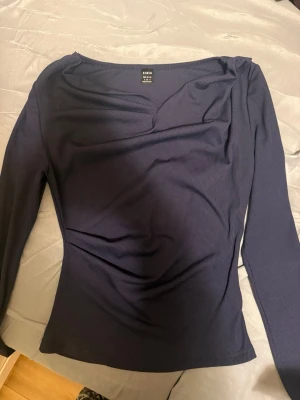 Mörkblå ribbad topp från Shein - Snygg mörkblå ribbad topp från Shein med lång ärm och lätt draperad halsringning. Toppen har en figurnära passform och är tillverkad i ett mjukt, stretchigt material som sitter skönt på kroppen. Perfekt att styla med jeans eller kjol.