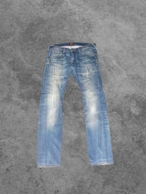 Blå slitna jeans från Lee - Säljer ett par blå jeans från Lee med snyggt slitna detaljer och ljusa partier framtill och baktill. Klassisk femficksmodell med raka ben och låg midja. Perfekta för en avslappnad och trendig look.