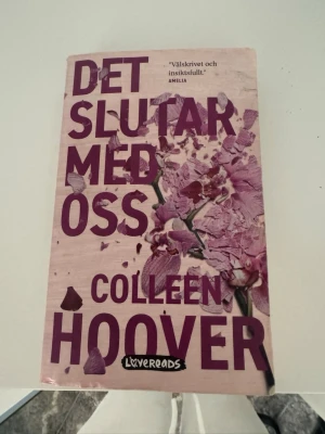 Det slutar med oss av Colleen Hoover - En gripande roman av Colleen Hoover med ett omslag i ljusrosa och lila toner, dekorerat med blommotiv. Boken är på svenska och ges ut av Lovereads. Perfekt för dig som gillar starka känslor och relationer i skönlitteratur.