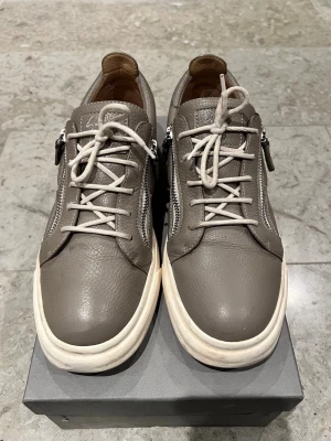 Grå sneakers från Giuseppe Zanotti - Snygga grå sneakers i läder från Giuseppe Zanotti med vita sulor, dragkedjor på sidorna och vita skosnören. Skorna har en stilren design med låg profil och detaljer i silver. Allt på bilderna medföljer. Säljer för att de inte passar längre. 