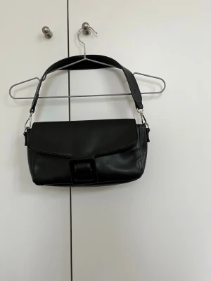 Svart axelväska med spänne - Snygg svart axelväska i slätt konstläder med bred axelrem och ett stort dekorativt spänne på framsidan. Väskan har en stilren, rektangulär form och silverfärgade metalldetaljer. Perfekt för dig som gillar minimalistisk och trendig design.