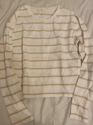 Gullig randig långärmad topp från Mango - Vit långärmad topp från Uniqlo med beigea horisontella ränder. Klassisk rak passform och rund halsringning. Tillverkad i mjuk bomull, perfekt för en avslappnad och stilren look. Katt finns i hemmet!
