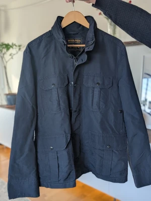 Mörkblå jacka från Woolrich nypris 6000! - Snygg mörkblå vindjacka från Woolrich med fyra stora fickor framtill och hög krage. Jackan har knappstängning och är långärmad. Tillverkad i slitstarkt material