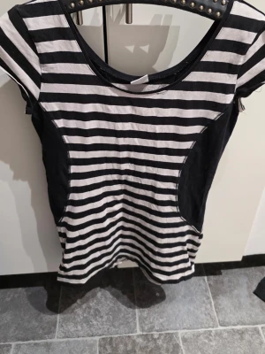 Randig svartvit topp i bomull - Säljer en svartvit randig topp med rund hals och korta ärmar. Toppen har breda svarta sidopaneler som ger en cool kontrast till de horisontella ränderna. Tillverkad i mjuk bomull som känns skön mot huden.