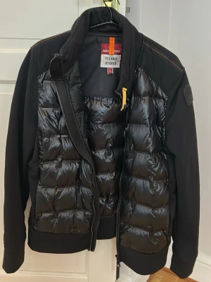 Svart pufferjacka från Parajumpers - Säljer en svart pufferjacka från Parajumpers med glansig, quiltad front och släta ärmar. Jackan har en gul detalj vid kragen och dragkedja framtill. Märket syns på ärmen och insidan har en tydlig Parajumpers Techno Hybrid-label. Perfekt för kalla dagar.