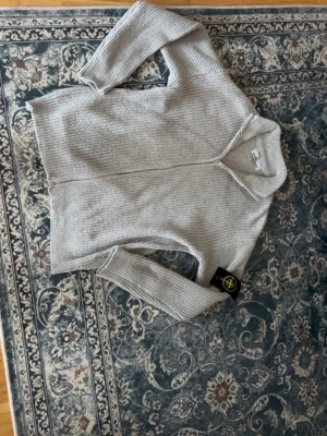 Ljusgrå ziptröja från Stone Island - Snygg ljusgrå stickad ziptröja från Stone Island med ribbade muddar och hög krage. Tröjan har dragkedja framtill och den klassiska Stone Island-loggan på ärmen. Perfekt för dig som gillar stilrena och exklusiva plagg.