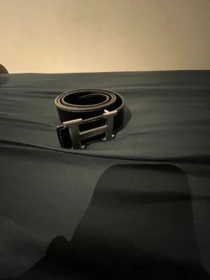 Svart skärp med H-spänne - Stilrent svart bälte med ett stort H-format metallspänne och vita kontrastsömmar längs kanterna. Bältet har en klassisk design som passar till många outfits och är tillverkat i skinn.