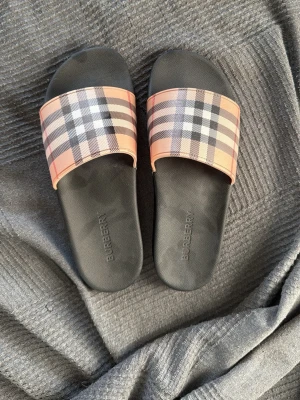 Burberry tofflor - Snygga tofflor från Burberry med klassiskt rutig design i beige, svart, vit och röd. Tofflorna har en bred rem över foten och svart sula med Burberry-logga. Perfekta för en trendig och avslappnan/sommar look. Pris går att diskutera vid snabb affär. 