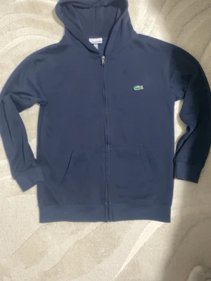 Mörkblå zip-hoodie från Lacoste - Mörkblå hoodie med dragkedja från Lacoste. Passar mellan S och M Klassisk krokodillogga på bröstet, huva och två stora fickor framtill. Mjuk bomullskänsla och ribbade muddar vid ärmslut och nederkant. Perfekt för en avslappnad och stilren look. Skickas samma dag pris kan diskuteras, 