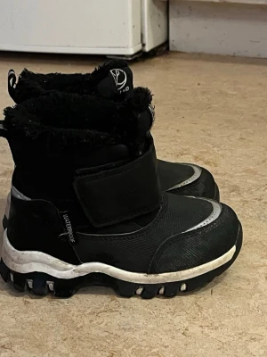 Svarta vinterkängor med kardborre - Svarta vinterkängor från Pax med grov sula och kardborreband. Kängorna är fodrade med mjukt, svart fuskpälsfoder och har en vit detalj runt sulan. Vattentäta och perfekta för kalla dagar. Reflekterande detalj framtill för extra synlighet.