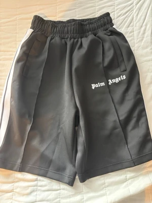 Palm Angels svarta shorts med vita ränder - Palm Angels shorts i svart med vita sidoränder och logga tryckt på vänster ben. De har elastisk midja och sidofickor. Perfekta för en avslappnad streetwear-look.