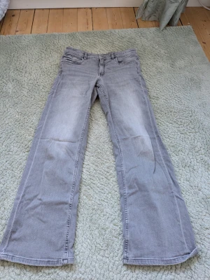 Grå bootcut jeans med fickdetaljer - Säljer ett par ljusgrå bootcut jeans med klassiska fem fickor, fram och bakfickor med lock och knapp. Jeansen har en avslappnad passform och är tillverkade i denim med lätt tvättad look. Perfekta för en casual stil.
