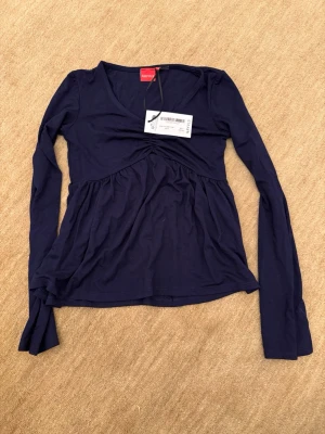 Mörkblå långärmad topp från Ryvls - Mörkblå långärmad Jersey topp från med v-ringning och lätt rynkad detalj under bysten. Tillverkad i mjuk bomull, perfekt för en avslappnad och stilren look. Passar dig som gillar enkel men snygg design.