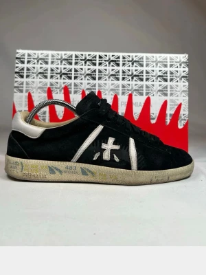 Svarta Premiata sneakers i mocka - Svarta sneakers från Premiata i mjuk mocka med vita detaljer och logga på sidan. Skorna har vita partier vid hälen och längs sidorna samt en beige sula med tryck. Klassisk låg modell med svarta skosnören och vadderad insida för extra komfort.