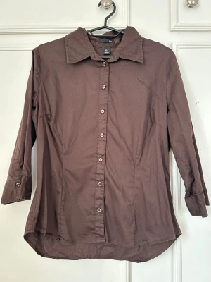 Brun skjorta med knappar - Brun skjorta i bomull med klassisk krage och långa ärmar. Skjortan har knappar framtill och vid ärmsluten, samt en något figursydd passform. Perfekt för en clean och enkel stil. OBS! Liten i storleken 