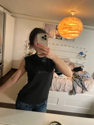 Svart ärmlös topp i glansigt material - Snygg svart ärmlös topp med rund halsringning. Toppen har en lätt glansig finish och en rak passform som sitter bekvämt.