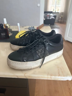 Svarta sneakers från tamaris - Svarta sneakers från tamaris med lågt skaft och vita sulor. Skorna har svarta snören och en snygg detalj i form av ett mönstrat glitter märke . Tillverkade i läderimitation med klassisk rund tå och bekväm passform. Tvättas såklart!