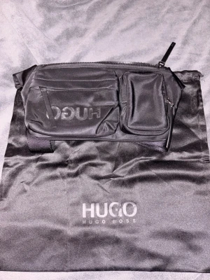 axelväska från Hugo Boss - Snygg svart magväska från Hugo Boss med flera fack och dragkedja. Väskan är tillverkad i ett slätt, tåligt material och har diskret logga framtill. Perfekt för att bära det viktigaste och hålla stilen. Levereras med original dustbag.