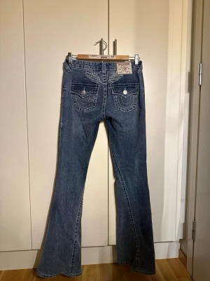 Blå bootcut jeans från True Religion - Säljer ett par blå bootcut jeans från True Religion med snygga kontrastsömmar och detaljerade bakfickor med lock och knappar. Jeansen har låg midja och klassisk femficksdesign. Perfekta för dig som gillar en retro vibe och vill sticka ut.