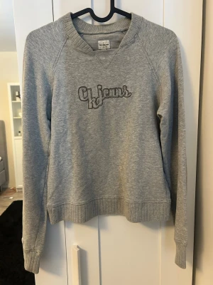 Grå sweatshirt från CK Jeans - Säljer en stilren grå sweatshirt från Calvin Klein Jeans med broderad logga på bröstet. Tröjan har rund halsringning, ribbade muddar och är tillverkad i mjukt bomullsmaterial. Perfekt för en avslappnad och trendig look.