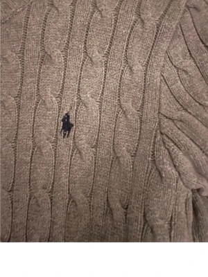 Kabelstickad tröja från Polo Ralph Lauren - Snygg kabelstickad tröja från Polo Ralph Lauren i en jordnära brungrå färg. Tröjan har rund halsringning och den klassiska broderade loggan på bröstet. Perfekt för dig som gillar tidlös och stilren design.