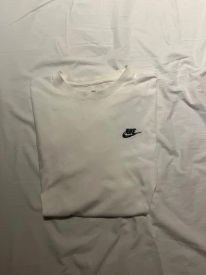 Vit t-shirt från Nike med logga - Klassisk vit t-shirt från Nike med liten svart logga på bröstet. T-shirten har rund hals och korta ärmar, perfekt för en clean och sportig look. Tillverkad i mjuk bomull för maximal komfort.