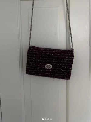 Mörklila axelväska med kedjerem - Snygg mörklila axelväska med silverfärgad kedjerem och twistlås framtill. Väskan har en rektangulär form och är gjord i ett stickat eller vävt material med diskreta vita detaljer. Perfekt för dig som vill ha en unik accessoar.
