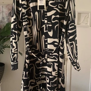 Svartvit mönstrad klänning från JDY - Cool svartvit klänning från JDY med abstrakt mönster, v-ringning och knytband i midjan. Långärmad modell med knappdetaljer vid ärmslut. Perfekt för dig som gillar statement-plagg och vill sticka ut med en unik look.
