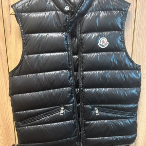 Moncler Gui Väst XS - Svart dunväst från Moncler med glansig finish och horisontella quiltade paneler. Västen har två snedställda fickor med dragkedja, hög krage med knapp och klassisk Moncler-logga på bröstet. Perfekt för lager-på-lager.