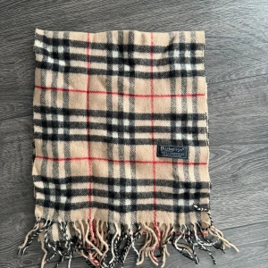 Rutig halsduk från Burberry - Snygg klassisk halsduk från Burberry i beige med svart, vit och röd rutig design. Gjord av 100% lammull och har fransar i ändarna. Perfekt accessoar för att lyfta din outfit med ikonisk brittisk stil.
