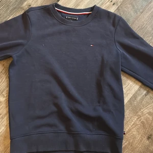 Mörkblå Tommy Hilfiger sweatshirt - Snygg mörkblå sweatshirt från Tommy Hilfiger med diskret logga på bröstet och tryck på ena ärmen. Tröjan har rund hals, ribbade muddar och är tillverkad i mjuk bomull. Perfekt för dig som gillar klassisk streetstil och vill ha något bekvämt och stilrent.