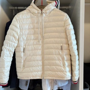 Vit Moncler dunjacka med huva - Snygg vit dunjacka från Moncler med huva och dragkedja framtill. Jackan har quiltad design, två fickor med dragkedja och Moncler-logga på ärmen. Röd, vit och marinblå rand på huva och ärmslut ger en sportig touch. Perfekt för kyliga dagar.