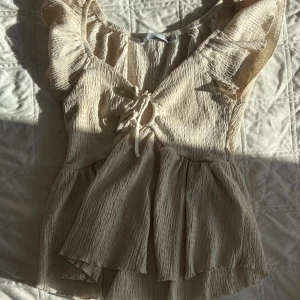 Beige omlottopp med volang från 157 - Supersöt beige omlottopp från 157 med volangdetaljer på ärmarna och v-ringning med knyt framtill. Toppens material är lätt skrynklat och ger en luftig känsla, perfekt för varmare dagar. Passformen är normal och toppen har korta ärmar.