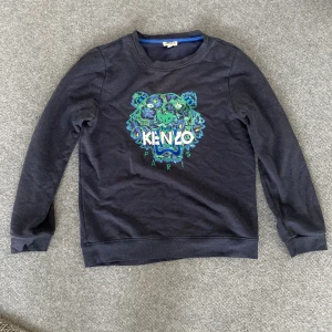 Svart Kenzo sweatshirt med tigertryck - Cool svart sweatshirt från Kenzo med ikoniskt tigertryck i grönt och blått framtill samt Kenzo Paris-logga. Tröjan har rund hals, långa ärmar och är gjord i mjuk bomull. Perfekt för dig som vill sticka ut med en streetstyle-vibe.