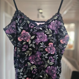 Blommig svart linnetopp med spets - Supersöt svart linnetopp från Hunkemöller med lila och rosa blommor, gröna blad och tunna axelband. Spetsdetaljer vid halsringningen ger en extra feminin touch. Perfekt att styla med jeans eller kjol för en chill look.