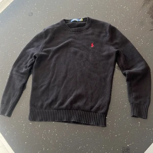 Svart stickad tröja Polo Ralph Lauren - Svart stickad tröja från Polo Ralph Lauren med rund halsringning och röd broderad logga på bröstet. Tröjan har ribbade muddar vid ärmslut och nederkant, perfekt för lager på lager under kyliga dagar.