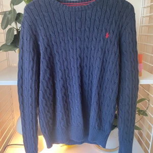 Mörkblå kabelstickad tröja Polo Ralph Lauren - Kabelstickad mörkblå tröja från Polo Ralph Lauren i storlek XL. Klassisk rund halsringning och röd broderad logga på bröstet. Tröjan är stickad i bomull och har långa ärmar med ribbade muddar för en snygg passform.