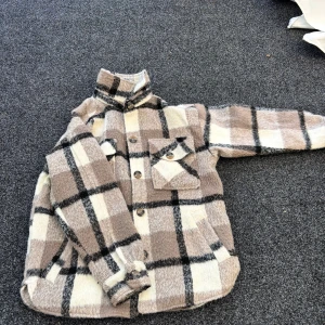 Rutig teddyjacka i beige och vitt - Mysig teddyjacka i beige, vitt och svart med rutigt mönster. Jackan har stora knappar framtill, bröstficka med lock och hög krage. Perfekt för dig som gillar en chill och trendig look.