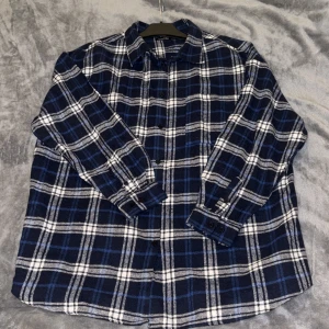 Blå och vit rutig skjorta från Boohoo - Snygg oversized rutig skjorta från Boohoo i mörkblått och vitt med inslag av ljusblått. Skjortan har klassisk krage, långa ärmar med manschett, två framfickor och knappar framtill. Perfekt för en chill och avslappnad stil. Materialet känns mjukt och bekvämt mot huden.
