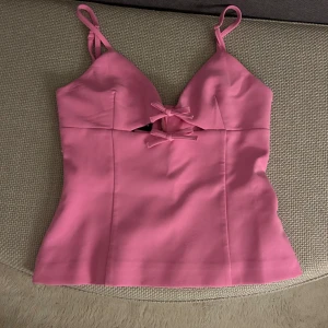 Rosa korsettopp med rosetter från Zara - Supersöt rosa korsettopp från Zara med smala axelband och två dekorativa rosetter framtill. Toppen har en snygg cut-out detalj under bysten och figurnära passform. Perfekt för dig som vill sticka ut med en feminin och trendig look.