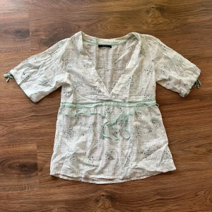 Vit omlott blus - Säljer en söt vit blommig omlottblus från Vero Moda i storlek M. Blusen har ljusgröna snörningsdetaljer i midjan och vid ärmslut, v-ringad hals och korta ärmar. Materialet är mjuk bomull med diskret rutmönster och små blommor i grönt och brunt.