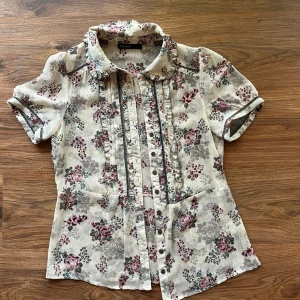 Blommig blus - Supersöt blommig skjorta från KappAhl i ljusa toner med rosa, grå och blå blommor. Skjortan har korta ärmar, volangdetaljer vid krage och knäppning framtill. Perfekt för dig som gillar romantisk och vintage-inspirerad stil. Har en liten fläck där fram, men annars är den i bra skick och inte använd så mycket☺️