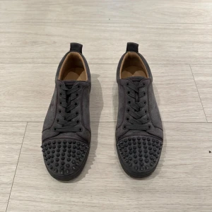 Christian louboutin - Snygga mörkgrå sneakers i mocka med rund tå och platt sula. Skorna har svarta snören och coola svarta nitar på tån för en edgy vibe. Insidan är beige och de har en låg profil som passar till streetwear. Perfekt för dig som gillar att sticka ut. Skorna är i nyskick och varsamt använda. Skriv vid frågor, Pris kan diskuteras vid snabb affär.