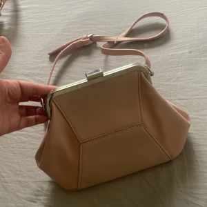 Ljusrosa handväska från David Jones - Snygg ljusrosa handväska från David Jones Paris i skinn med silverfärgade metalldetaljer och kedja. Väskan har en unik geometrisk form, metallram och justerbar axelrem. Perfekt för dig som gillar stilrena och moderna accessoarer.