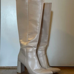 Beiga höga stövlar med klack - Snygga höga stövlar i beige skinnimitation med spetsig tå och bred klack. Stövlarna har en stilren och minimalistisk design som ger en cool vibe. Perfekta för dig som vill sticka ut med ett par statement boots i ljus ton. Storlek 40 med 9 cm klack. 😚