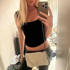 Vit/grå axelväska med kedja från Stella Mccartney - Säljer en vit/grå axelväska i mocka från Stella Mccartney med snygga kedjedetaljer runt kanterna och som axelrem. Väskan har en rektangulär form och stängs med magnetlås. Perfekt storlek för det viktigaste och ger en cool touch till din outfit. Väskan har en liten fläck på framsidan, inte provat att tvätta bort den.