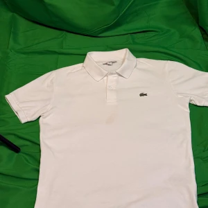 Vit pikétröja  Lacoste - Klassisk vit pikétröja  Lacoste med den ikoniska gröna krokodilloggan på bröstet. Tröjan har korta ärmar, krage och knäppning med två knappar framtill. Tillverkad i mjuk bomull som känns skön mot huden. Perfekt för en clean och stilren look.Liknande modell som originalet!
