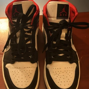 Nike Air Jordan 1 svart/vit/röd 39 - Säljer ett par Nike Air Jordan 1 i klassisk svart, vit och röd färg. Skorna har högt skaft, svart swoosh och röda detaljer vid ankeln. Tillverkade i skinn med perforerad tå och svarta skosnören. Perfekta för dig som vill sticka ut med en ikonisk sneaker.
