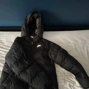 Svart pufferjacka från Nike med huva - Svart pufferjacka från Nike med huva och dragkedja framtill. Jackan har en clean look med vit Nike-logga på bröstet och är tillverkad i syntetmaterial. Perfekt för små barn, kalla dagar och har en skön, vadderad känsla.